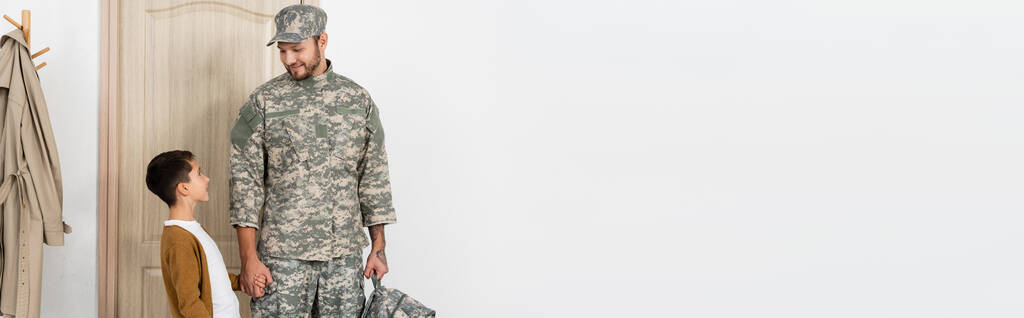 Στρατιωτικός άντρας με σακίδιο να κρατάει το χέρι του γιου του να τον συναντήσει κοντά στην πόρτα στο σπίτι, πανό - Φωτογραφία, εικόνα