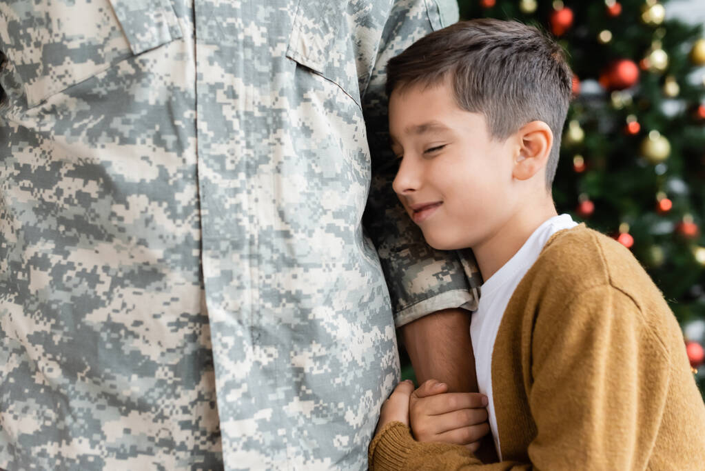 mosolygós fiú csukott szemmel öleli az apa kezét katonai egyenruhában  - Fotó, kép