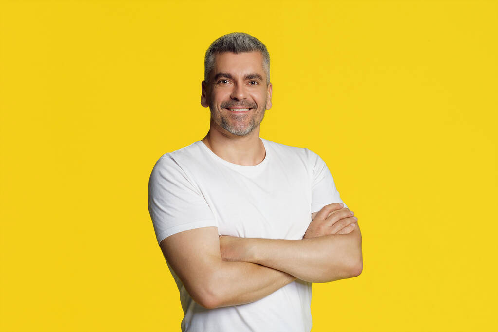 Feliz sonriente hombre atlético de pelo gris de mediana edad posando con las manos dobladas usando una camiseta blanca aislada sobre fondo amarillo. Ajuste reflexivo, hombre musculoso de mediana edad. - Foto, Imagen