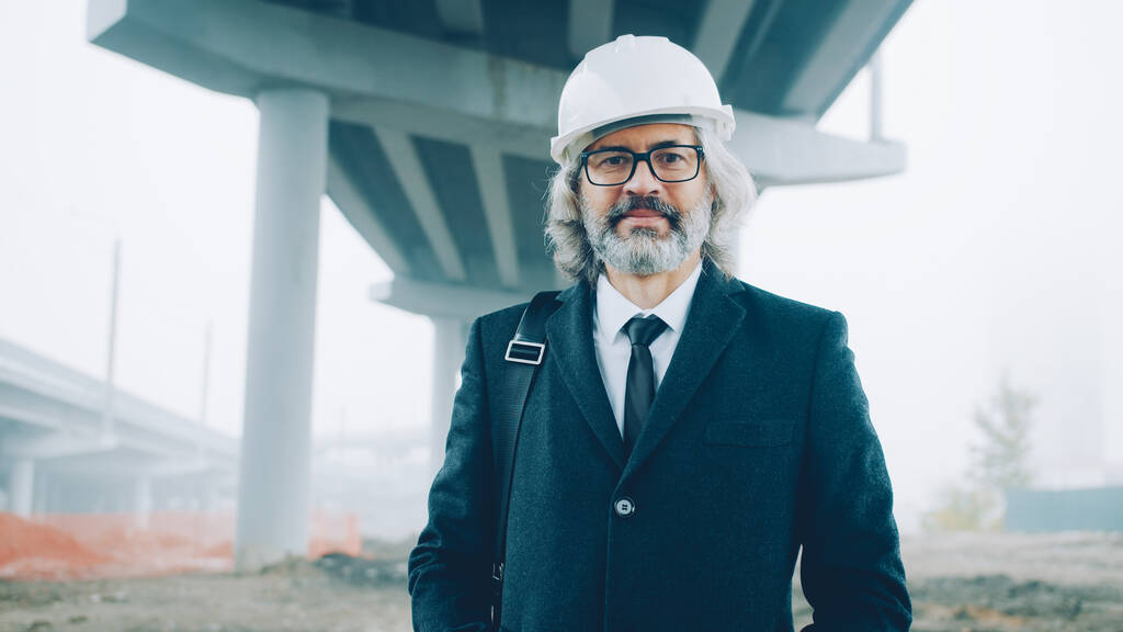 Portrait d'un homme architecte prospère debout à l'extérieur sur le chantier portant un casque de sécurité et une combinaison formelle et regardant la caméra. - Photo, image