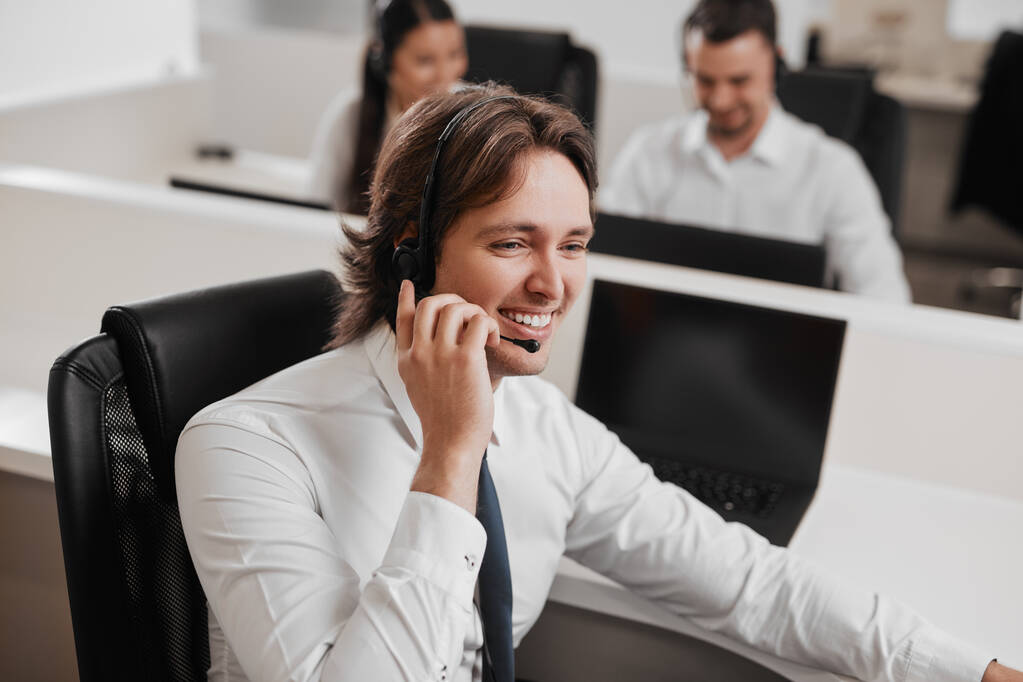 Alto ángulo de hombre alegre en desgaste formal sonriendo y hablando con el cliente a través de auriculares durante el trabajo en la oficina de línea de ayuda - Foto, imagen