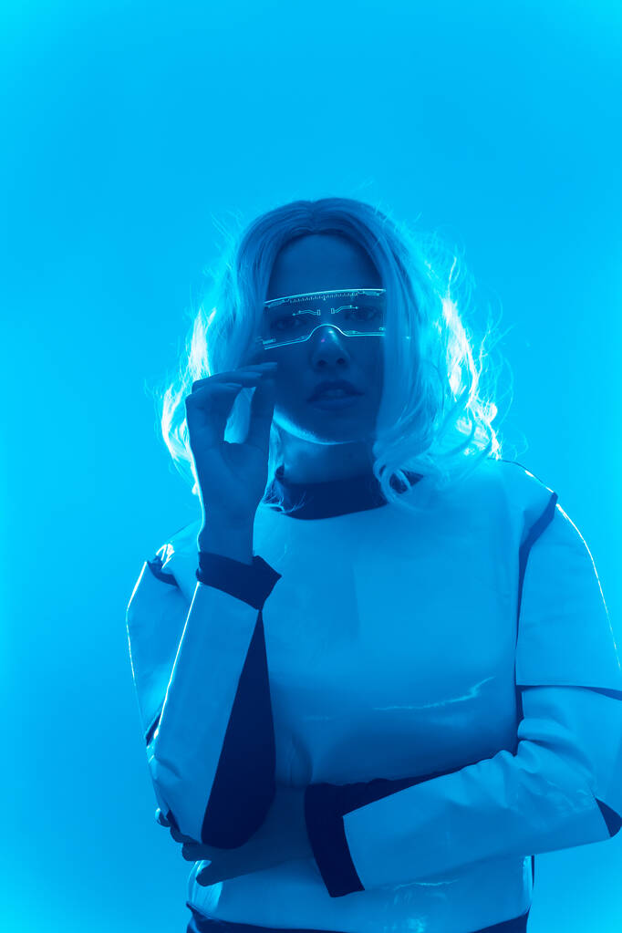 Jeune femme avec costume futuriste et lunettes avec lumières LED bleues, concept virtuel ou métaverse, regardant la caméra - Photo, image