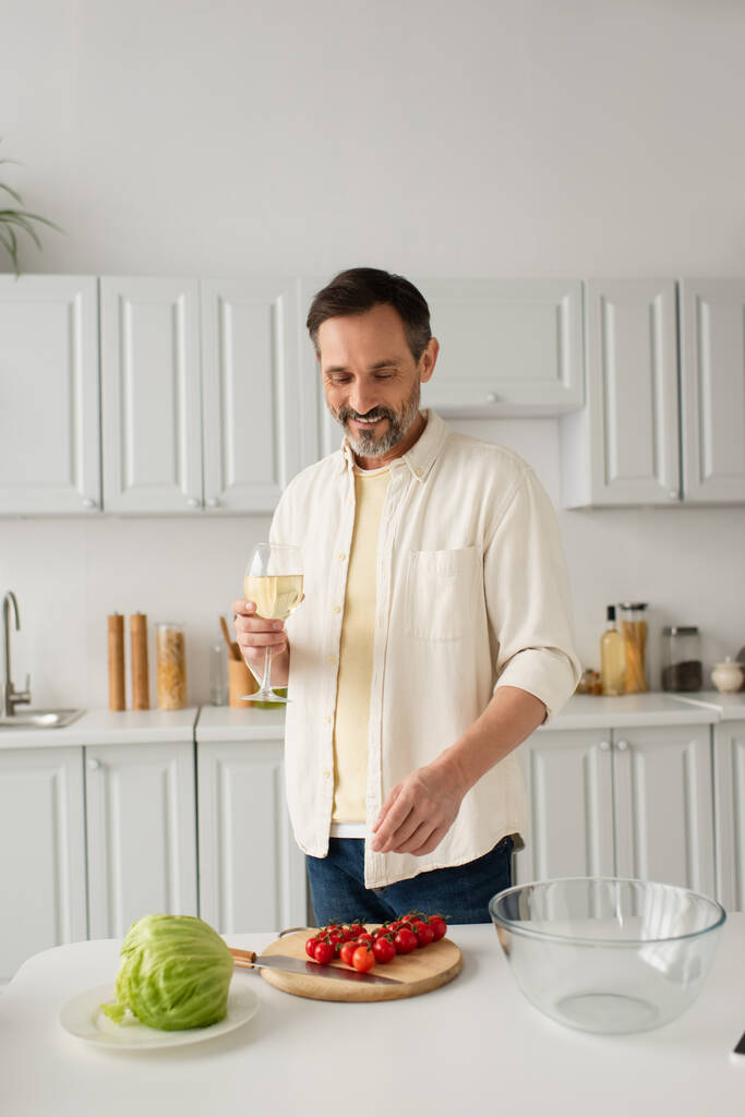 hombre alegre en camisa blanca sosteniendo copa de vino cerca de lechuga fresca y tomates cherry maduros - Foto, imagen