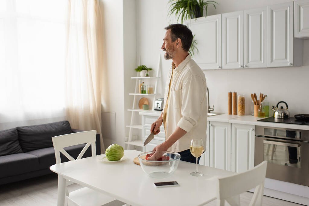 szczęśliwy brodaty mężczyzna patrząc w pobliżu świeżych warzyw i smartfona z pustym ekranem w kuchni - Zdjęcie, obraz