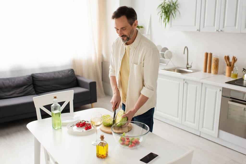 hombre sonriente cortando lechuga cerca del tazón con ensalada de verduras y botellas con aceite - Foto, Imagen
