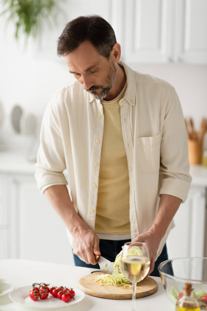 γενειοφόρος άνδρας σε λευκό πουκάμισο κοπής μαρούλι κοντά ώριμα ντοματίνια και θολή ποτήρι κρασί - Φωτογραφία, εικόνα