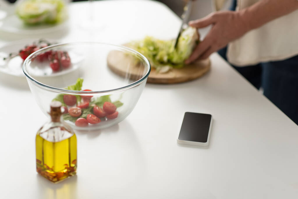 обрезанный вид размытого человека режет салат при приготовлении салата рядом с сотовым телефоном с пустым экраном и бутылкой масла - Фото, изображение