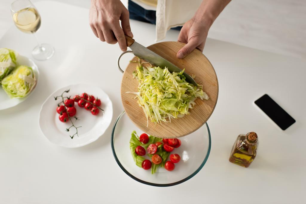 вид сверху на обрезанного человека, добавляющего салат в миску с помидорами черри возле смартфона с пустым экраном  - Фото, изображение