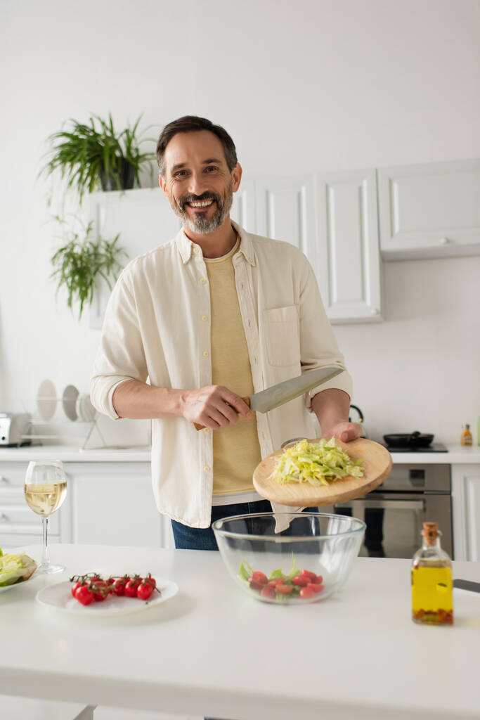 uomo barbuto preparare insalata con lattuga e pomodorini mentre sorride alla macchina fotografica - Foto, immagini
