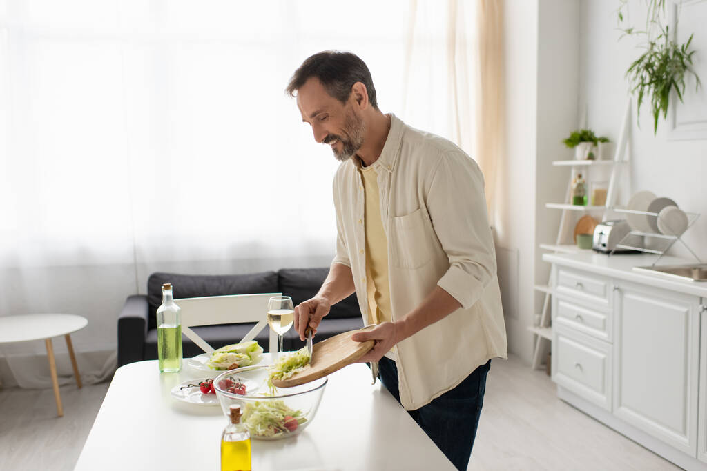 счастливый человек держит разделочную доску рядом с миской во время приготовления овощного салата с салатом и помидорами черри - Фото, изображение