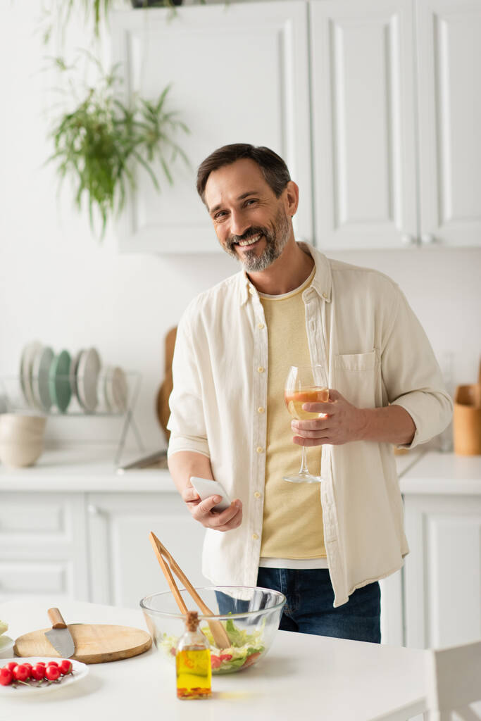 fröhlicher Mann mit Handy und Glas Wein, der in die Kamera neben Schüssel mit Salat und Flasche Öl blickt - Foto, Bild