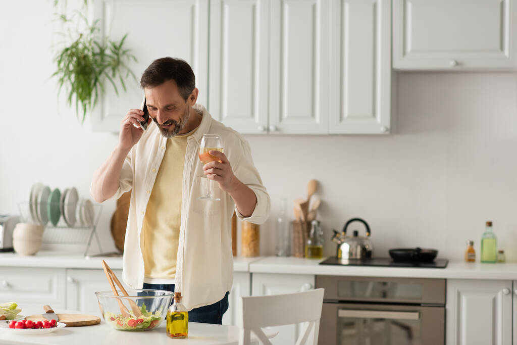 Lächelnder Mann mit Weinglas im Smartphone-Gespräch in der Nähe von Kirschtomaten und Schüssel mit Salat - Foto, Bild