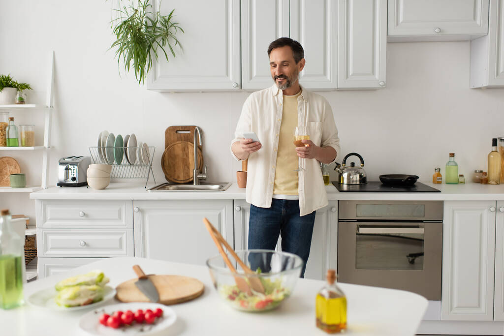 sonriente hombre con copa de vino usando teléfono inteligente cerca de verduras frescas y vasos con aceite en primer plano borroso - Foto, Imagen