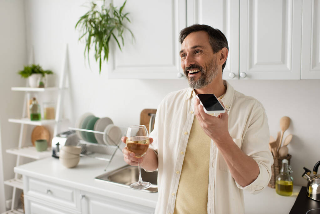 весёлый мужчина с бокалом белого вина и отправкой голосового сообщения на сотовый телефон на кухне - Фото, изображение