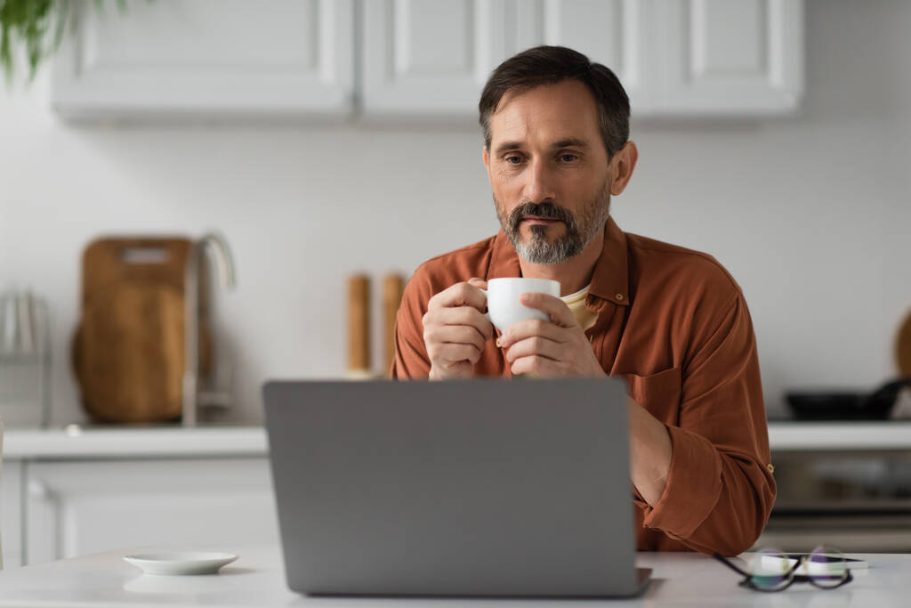 задумчивый бородатый мужчина держит чашку кофе и смотрит на ноутбук на кухне - Фото, изображение