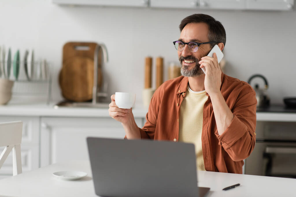 Gözlüklü neşeli bir adam kahve fincanı tutuyor ve cep telefonuyla konuşuyor. Bulanık dizüstü bilgisayarın yanında. - Fotoğraf, Görsel