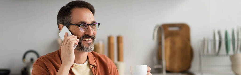 hombre sonriente en anteojos sosteniendo taza de café y hablando por teléfono celular en la cocina, pancarta - Foto, Imagen