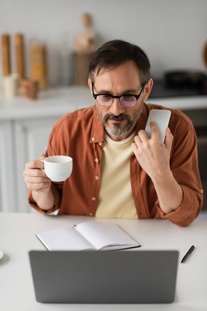 Gözlüklü adam elinde akıllı telefon ve kahve fincanı tutarken boş defterin yanındaki bulanık dizüstü bilgisayara bakıyor. - Fotoğraf, Görsel