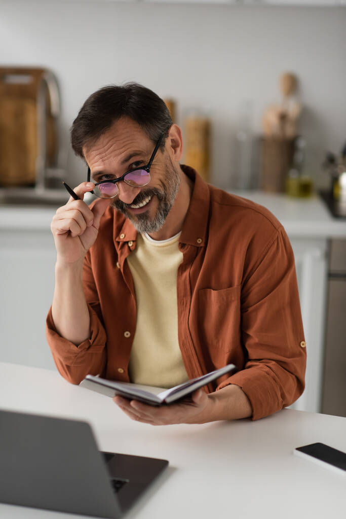 fröhlicher Mann mit Brille hält Notizbuch und Stift in der Hand und lächelt in die Kamera neben verschwommenem Laptop - Foto, Bild