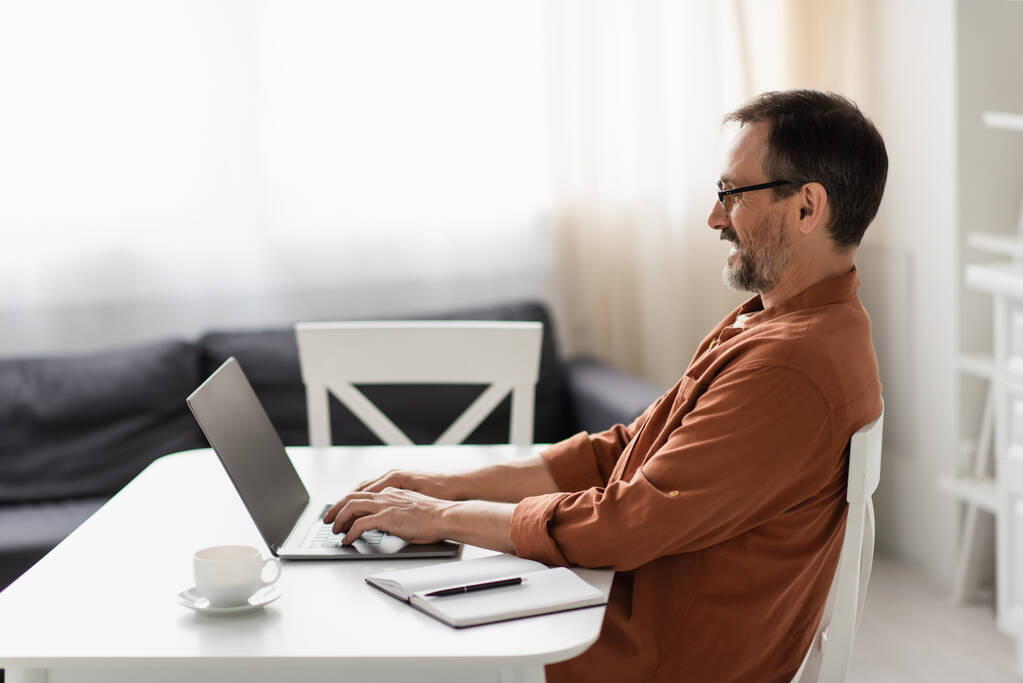 vista laterale di uomo felice in occhiali da vista digitando sul computer portatile con schermo vuoto vicino notebook vuoto e tazza di caffè - Foto, immagini