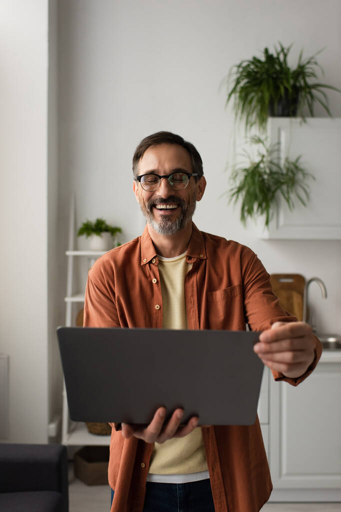 izgatott férfi szemüvegben tartja laptop és nevetés csukott szemmel a konyhában - Fotó, kép