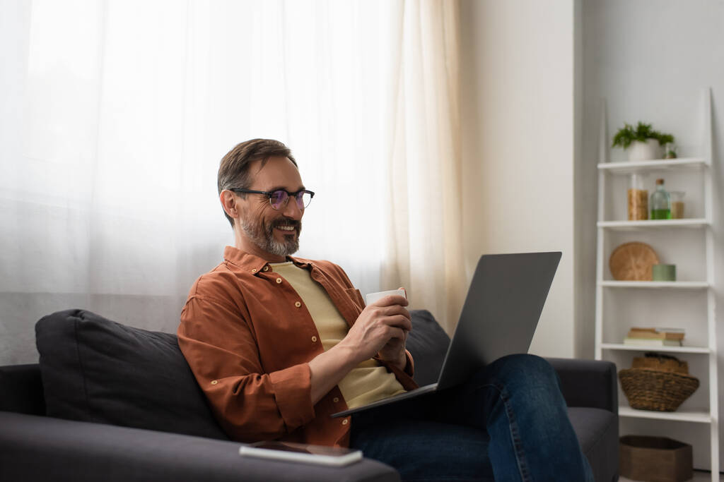 cheerful man in eyeglasses sitting on couch with coffee cup and laptop - Photo, Image