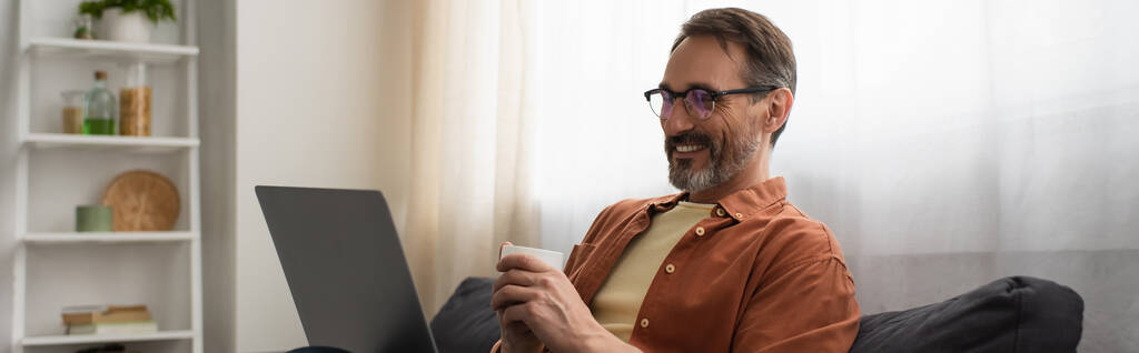眼鏡をかけた陽気な男がコーヒーカップを持ってノートパソコンやバナーを見て - 写真・画像