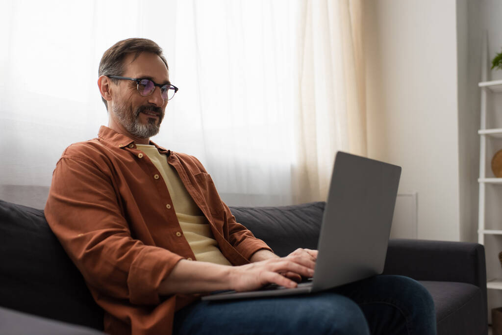позитивный бородатый мужчина в очках сидит на диване и печатает на ноутбуке - Фото, изображение