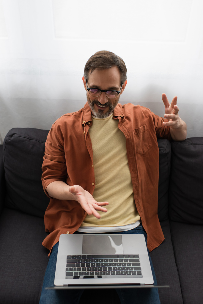 自宅のソファでビデオチャット中にノートパソコンを指している眼鏡の笑顔の男の高い角度のビュー - 写真・画像