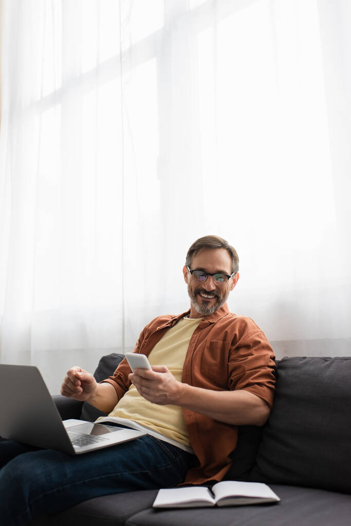 sonriente hombre barbudo sosteniendo el teléfono inteligente y mirando el cuaderno vacío mientras está sentado en el sofá con el ordenador portátil - Foto, imagen