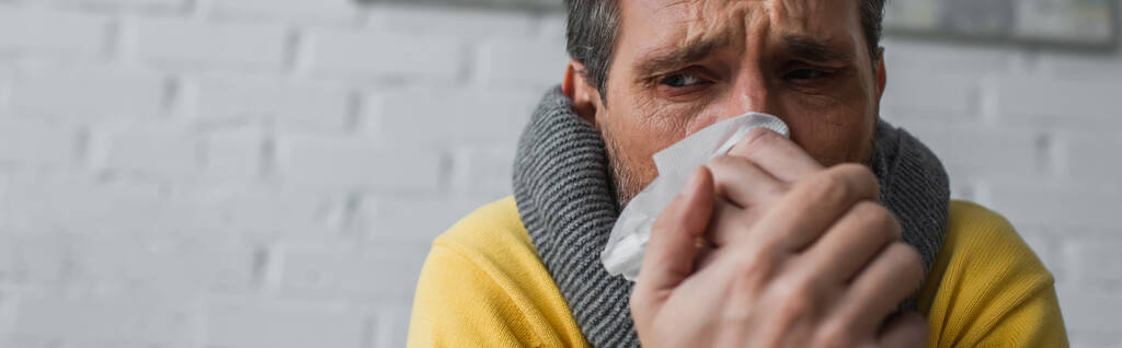 ill man in warm scarf suffering from runny nose and holding paper napkin near face, banner - Photo, Image
