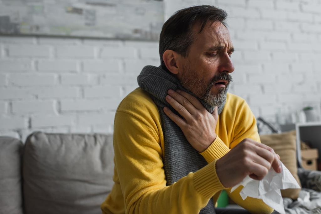 άρρωστος άνθρωπος σε ζεστό κασκόλ αγγίζοντας πονόλαιμο και βήχα κρατώντας χαρτοπετσέτα - Φωτογραφία, εικόνα