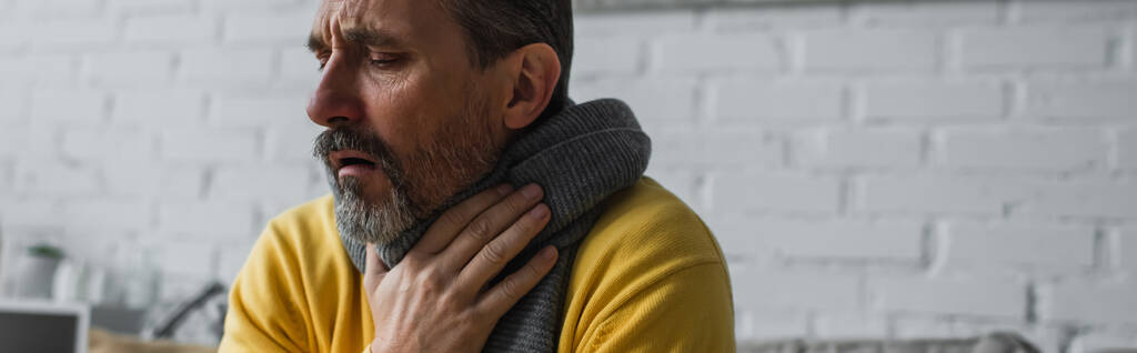 暖かいスカーフに身を包んだ病気の男が喉を痛めて家で咳をしバナー - 写真・画像
