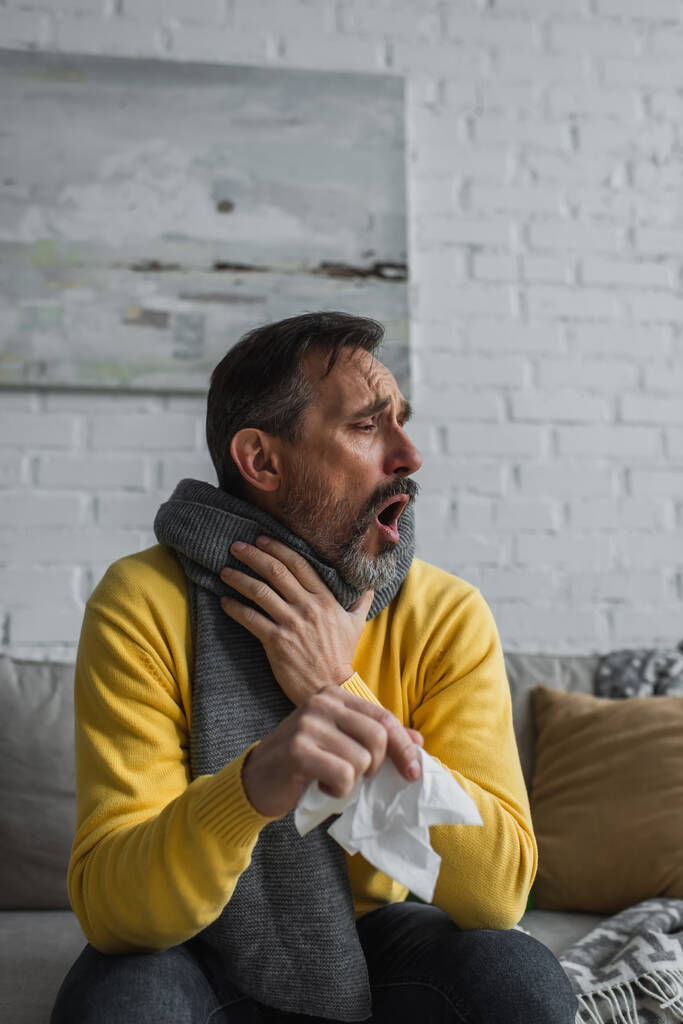 άρρωστος άνδρας με ζεστό μαντήλι κρατώντας χαρτοπετσέτα και βήχοντας στον καναπέ - Φωτογραφία, εικόνα