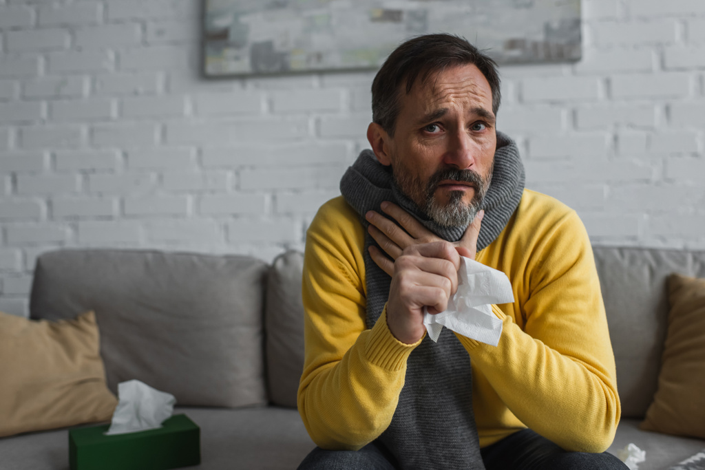 хворий чоловік в теплому шарфі торкається болю в горлі, тримаючи паперову серветку і дивлячись на камеру
 - Фото, зображення