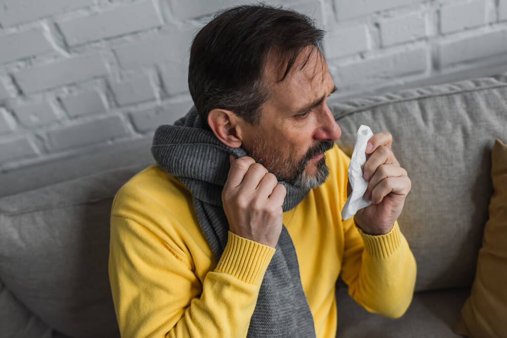больной человек в теплом шарфе держит бумажную салфетку, сидя дома на диване - Фото, изображение