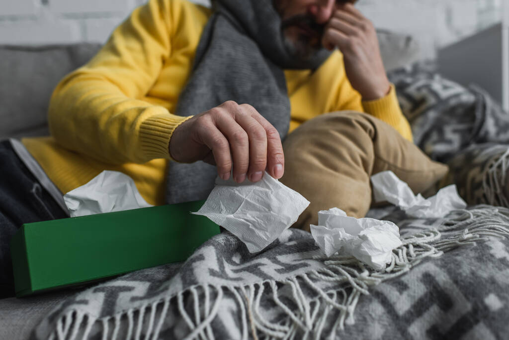 частичный вид больного, лежащего на диване с теплым одеялом и держащего бумажную салфетку - Фото, изображение