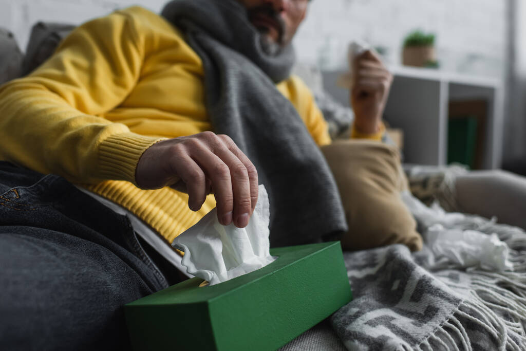 Teilbild eines kranken Mannes, der auf Couch liegt und Papierserviette aus Packung nimmt - Foto, Bild