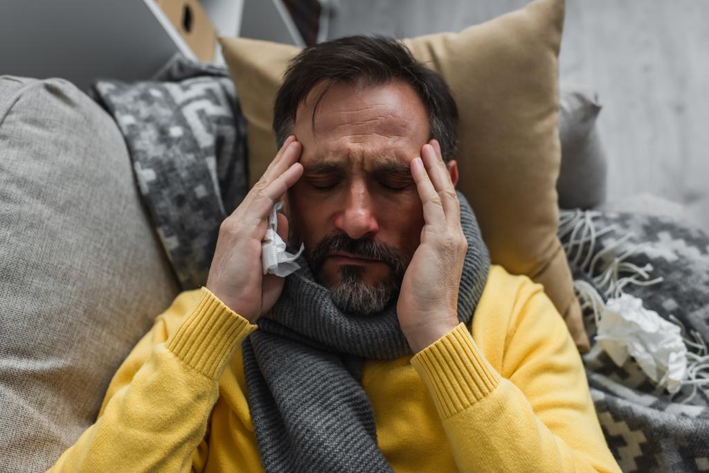 vista superior del hombre enfermo en una cálida bufanda tocando la cabeza mientras está acostado en el sofá y sufre de migraña - Foto, Imagen