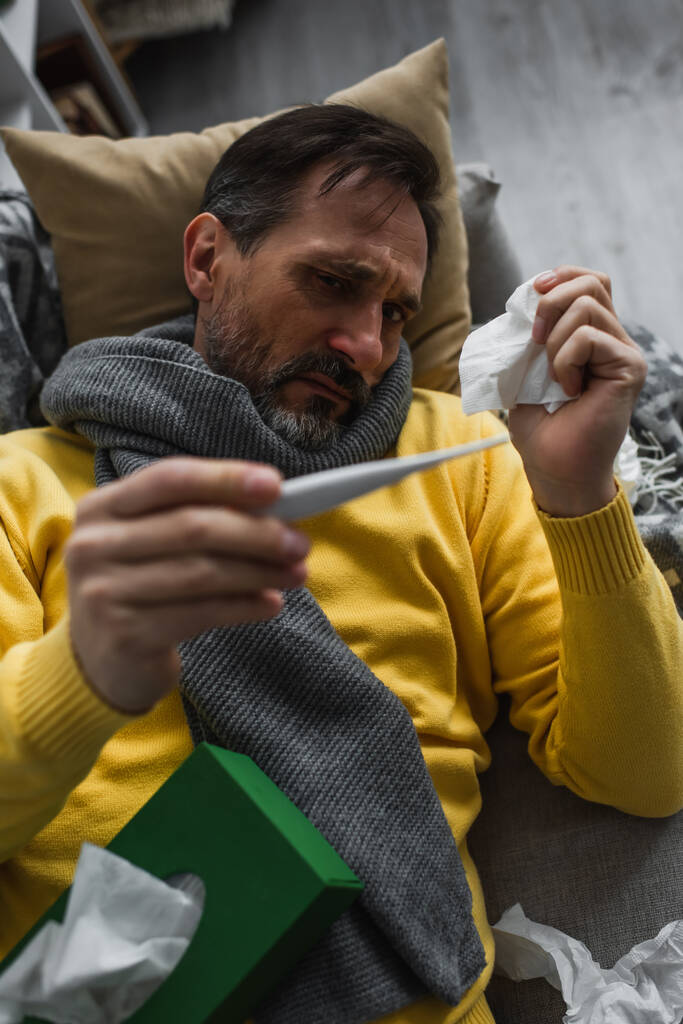хворий чоловік в теплому шарфі тримає паперову серветку і дивиться на електронний термометр
 - Фото, зображення