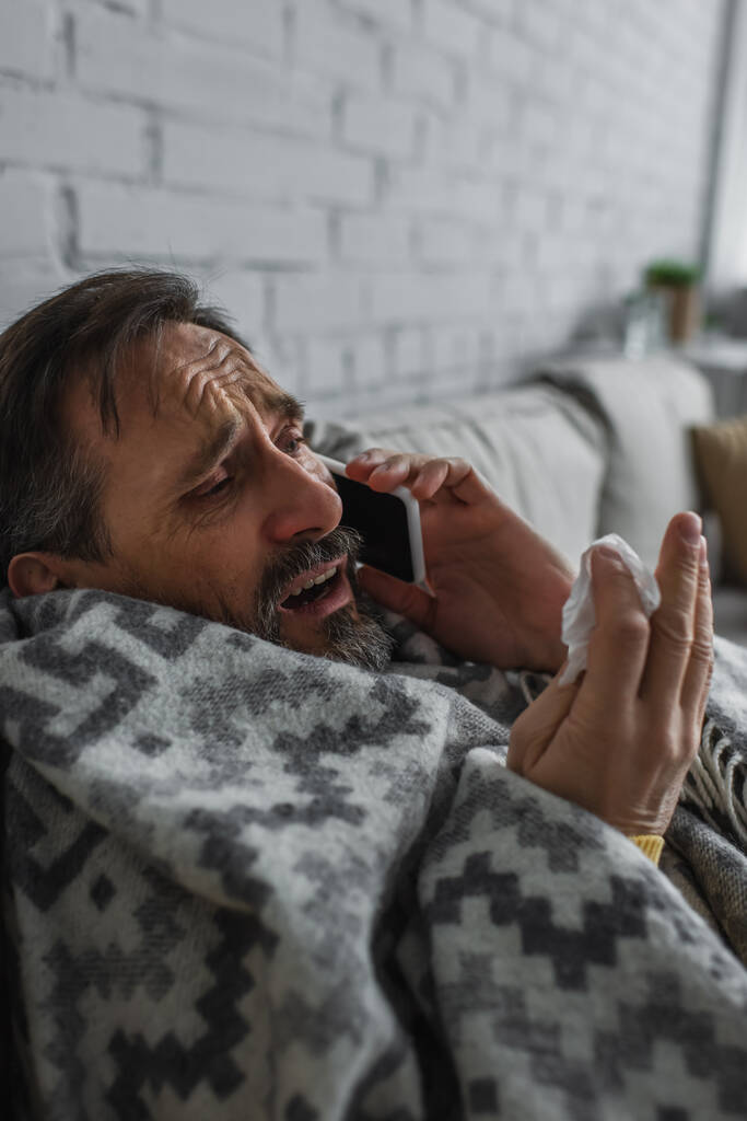 homem doente envolto em cobertor quente segurando guardanapo de papel e falando no telefone móvel - Foto, Imagem