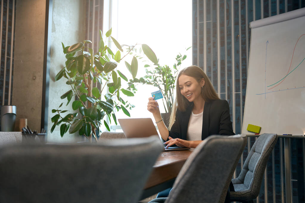 Працівник компанії сидить за офісним столом з дебетовою карткою в руці і друкує на ноутбуці
 - Фото, зображення
