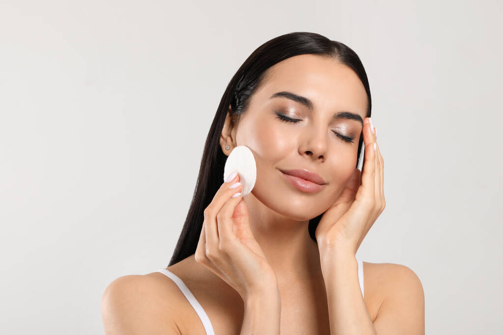 Young woman using cotton pad with micellar water on light grey background - Foto, afbeelding