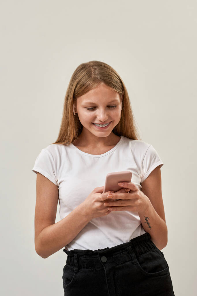 Gericht Kaukasisch tienermeisje draagt oortjes browsen smartphone. Mooi blond vrouwelijk kind van zoomer generatie. Moderne jonge levensstijl. Geïsoleerd op witte achtergrond in de studio. Kopieerruimte - Foto, afbeelding