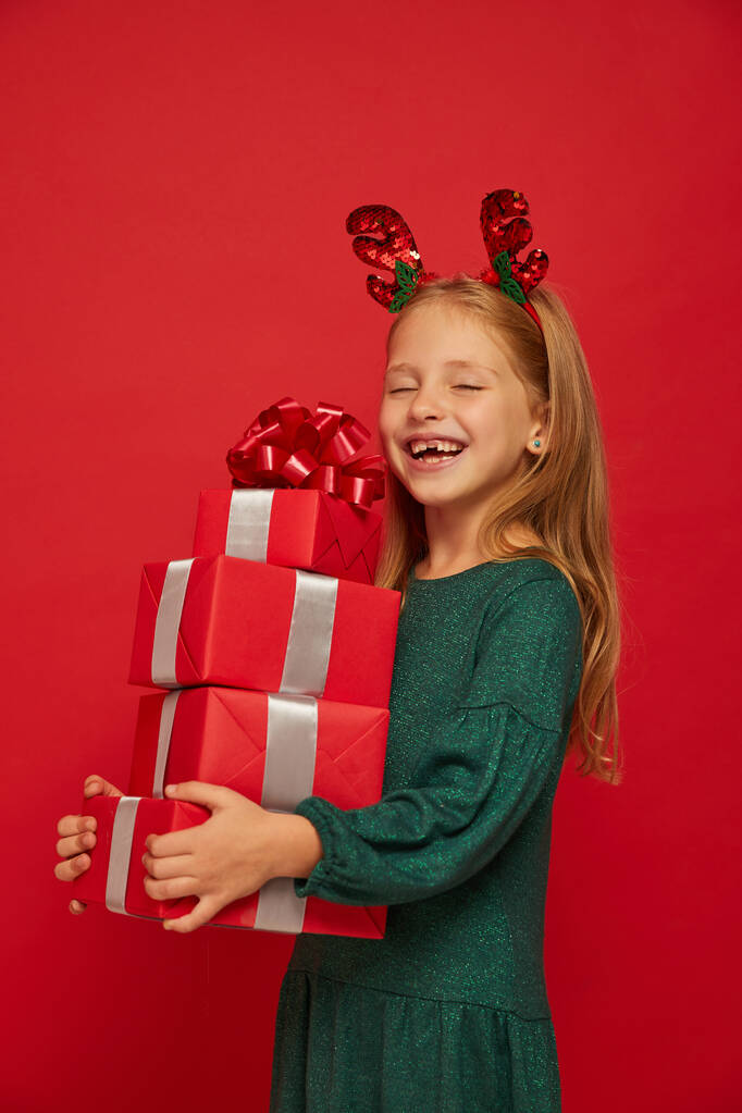Enfant drôle souriant (enfant, fille) dans un bandeau de rennes. Tenir les cadeaux de Noël à la main. Concept de Noël. Tir sur fond rouge - Photo, image