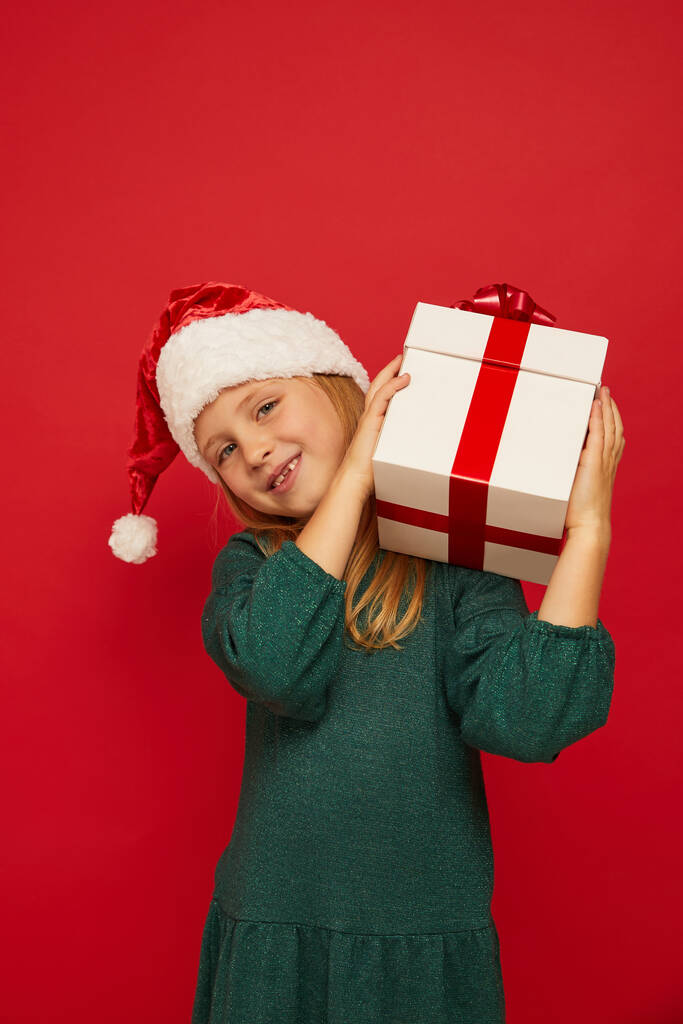 Enfant drôle souriant (enfant, fille) dans le chapeau rouge Santa tenant boîte cadeau de Noël à la main. Concept de Noël. Tir sur le fond rouge - Photo, image