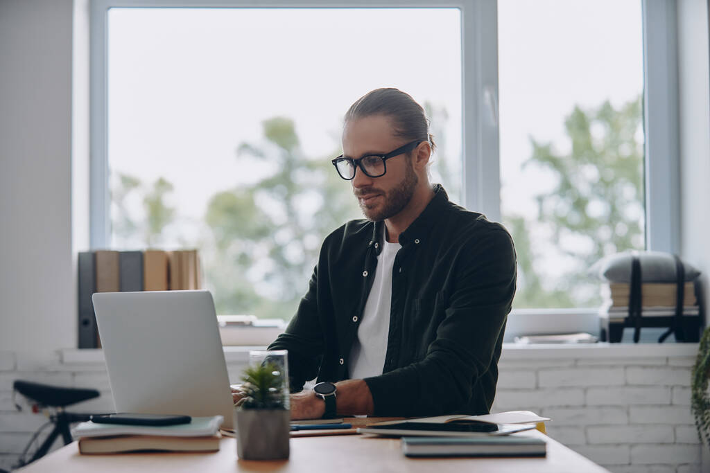 Jeune homme confiant utilisant un ordinateur portable tout en étant assis à son lieu de travail dans le bureau - Photo, image