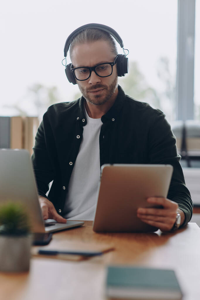 Впевнена людина в навушниках, використовуючи технології, сидячи на своєму робочому місці в офісі
 - Фото, зображення