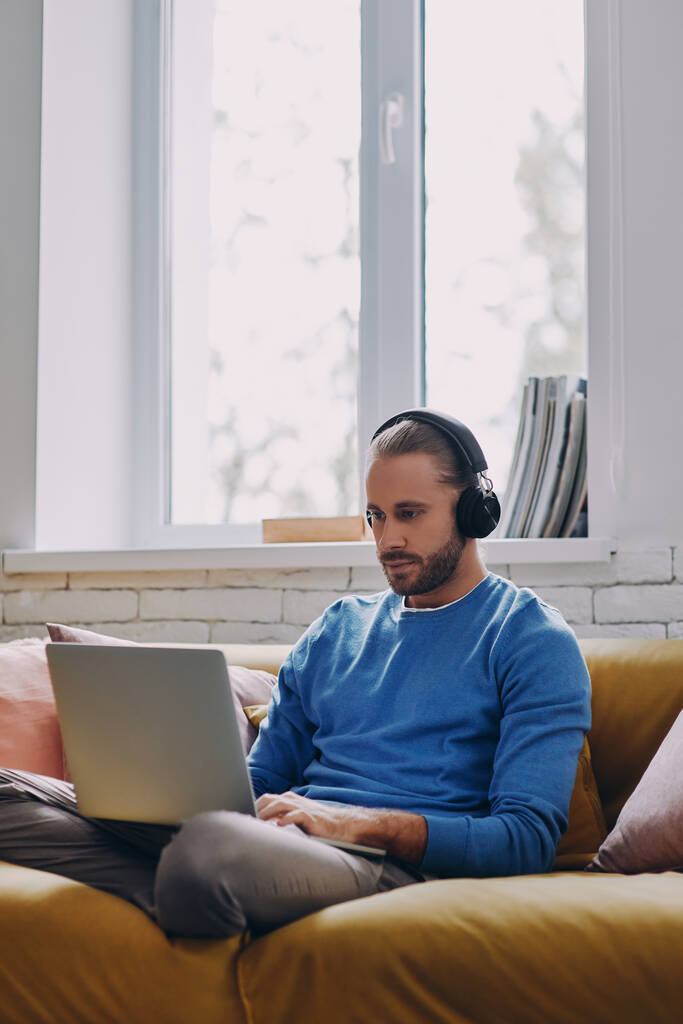 Schöner junger Mann mit Kopfhörern arbeitet am Laptop, während er zu Hause auf der Couch sitzt - Foto, Bild