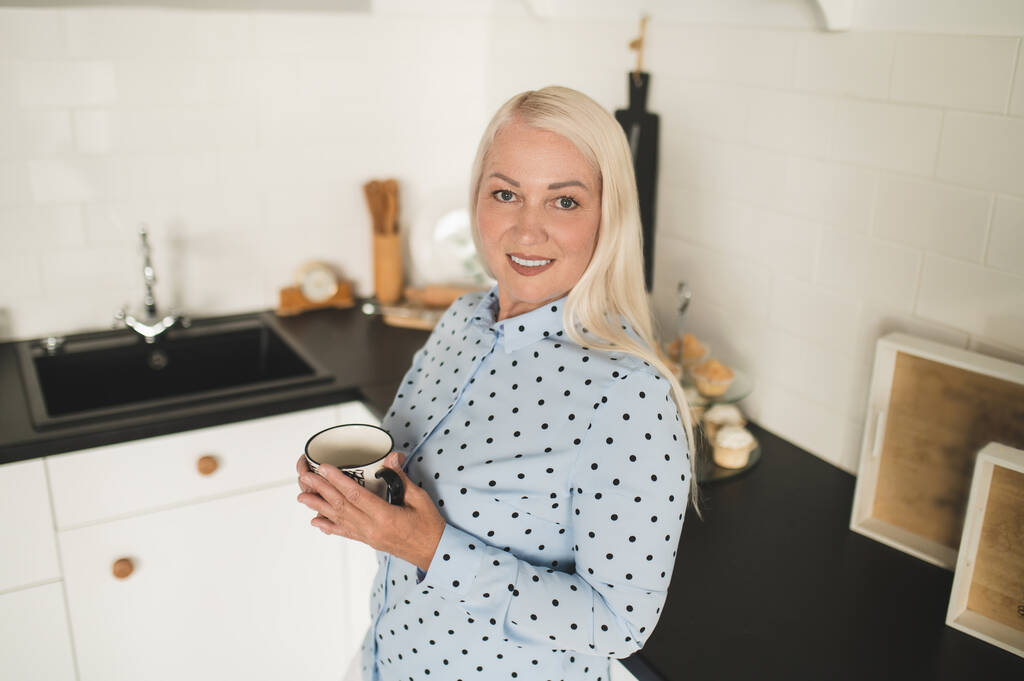 Sorridente piacere donna dai capelli lunghi con una tazza di ceramica tra le mani appoggiata al bancone della cucina - Foto, immagini